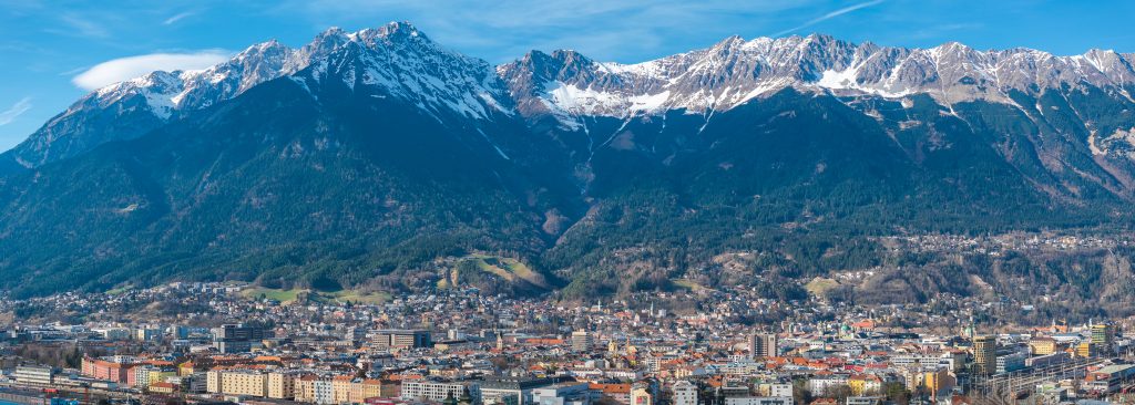 Aerial view of Innsbruck, Austria, featuring colorful buildings, historic architecture, and the snow capped Nordkette mountain range under a clear blue sky.